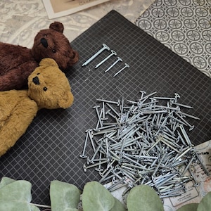 May include: A collection of small, silver nails and four larger silver tacks are arranged on a black grid surface. Two plush teddy bears in brown and tan are positioned on the left side of the image, suggesting a craft or DIY project.