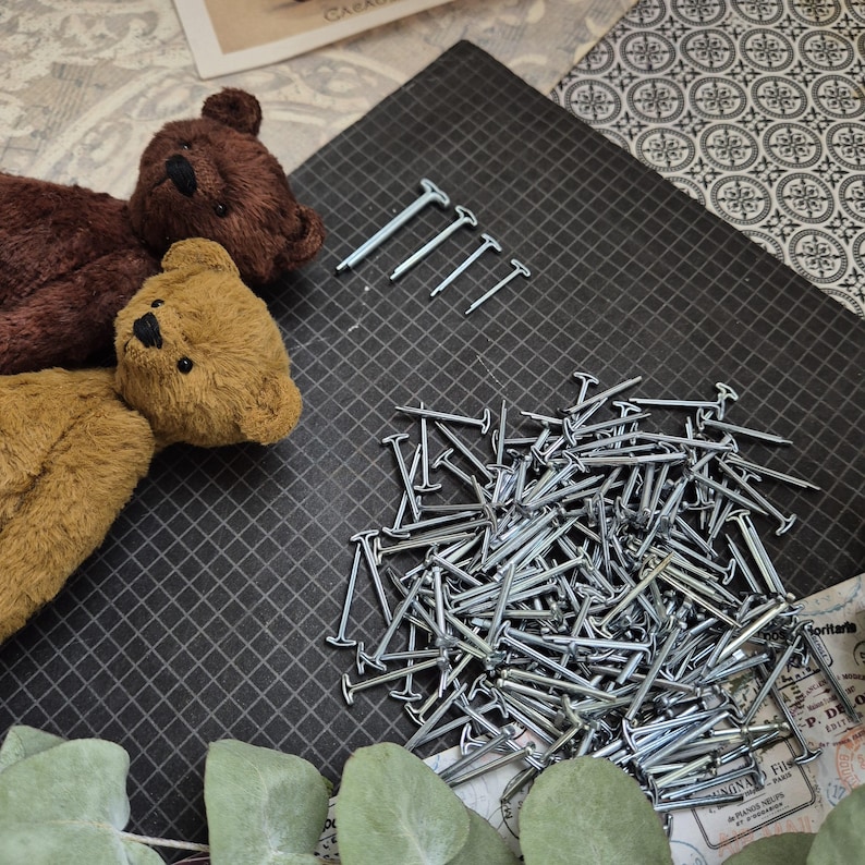 May include: A collection of small, silver nails and four larger silver tacks are arranged on a black grid surface. Two plush teddy bears in brown and tan are positioned on the left side of the image, suggesting a craft or DIY project.
