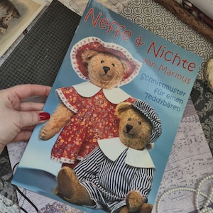 Op de afbeelding: Een softcover boek getiteld Neffe & Nichte von Marinus met patronen voor teddyberen. De kaft toont twee lichtbruine teddyberen, één in een rode bloemetjesjurk met een witte kraag en één in een blauw gestreepte outfit.