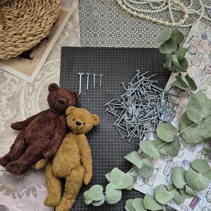 May include: Two handmade teddy bears, one brown and one tan, rest on a black grid surface next to a pile of nails and a few individual nails. Eucalyptus leaves and decorative paper complete the scene.