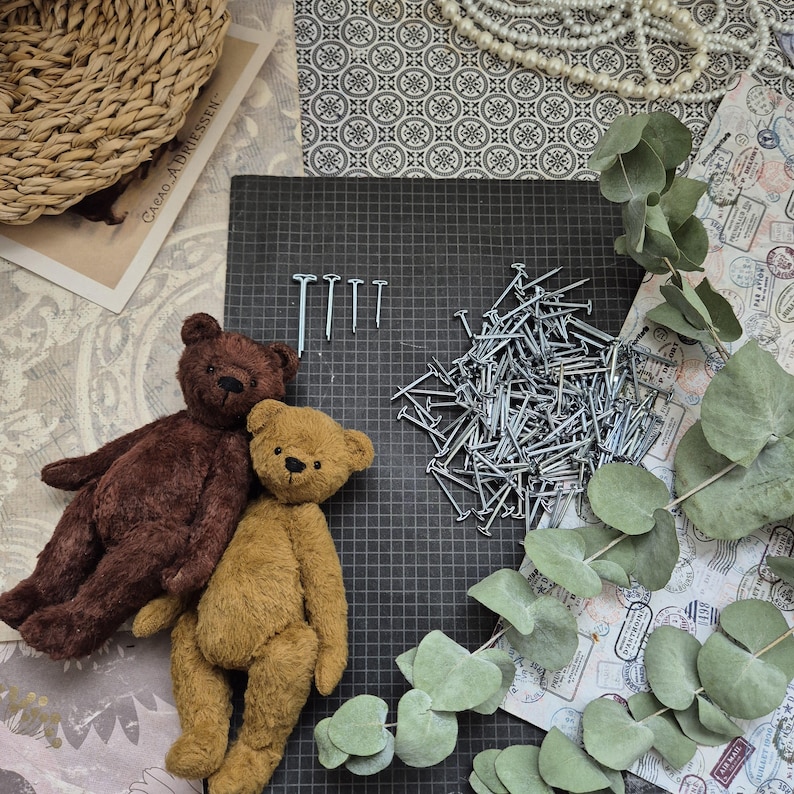 May include: Two handmade teddy bears, one brown and one tan, rest on a black grid surface next to a pile of nails and a few individual nails. Eucalyptus leaves and decorative paper complete the scene.