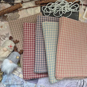 Op de afbeelding: Een verzameling gevouwen stoffen vierkanten in verschillende kleuren, waaronder rood, blauw en bruin, met een klassiek ruitpatroon. De stoffen zijn gerangschikt op een oppervlak met decoratieve elementen zoals houten takjes, kralen en kleine kabouterfiguren.
