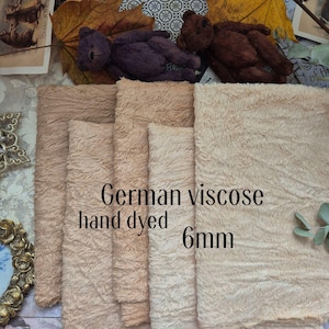 Può includere: Primo piano di diversi pezzi di tessuto in viscosa tedesca tinto a mano in varie tonalità di beige e marrone. Il tessuto ha un aspetto strutturato e morbido. Il testo sul tessuto recita "German viscose hand dyed 6mm". Due piccoli orsacchiotti sono visibili sullo sfondo.