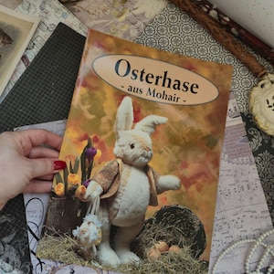 May include: A rectangular book titled Osterhase - aus Mohair - featuring a cream-colored plush rabbit wearing a brown patterned vest. The rabbit stands near a dark basket filled with hay and eggs, set against a warm, autumn-toned background.