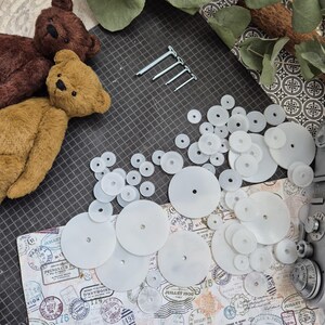 Op de afbeelding: Bovenaanzicht van teddybeerbenodigdheden. Verschillende kleine, bruine teddyberen zijn te zien, samen met witte plastic schijven en metalen bevestigingsmiddelen. De items zijn gerangschikt op een patroon, wat duidt op een knutselproject.