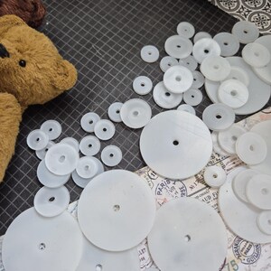 Op de afbeelding: Een verzameling witte plastic schijven met centrale gaten, verspreid op een donker oppervlak. De schijven vari&euml;ren in grootte en dikte, wat suggereert dat het componenten zijn voor een knutsel- of speelgoedproject. Een deel van een bruine teddybeer is zichtbaar.