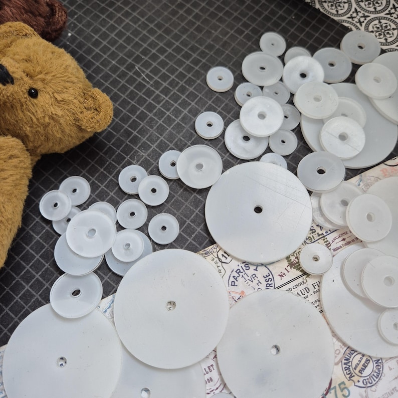 Op de afbeelding: Een verzameling witte plastic schijven met centrale gaten, verspreid op een donker oppervlak. De schijven vari&euml;ren in grootte en dikte, wat suggereert dat het componenten zijn voor een knutsel- of speelgoedproject. Een deel van een bruine teddybeer is zichtbaar.