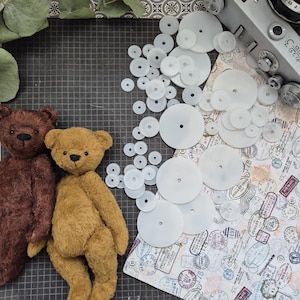 Op de afbeelding: Twee handgemaakte teddyberen, &eacute;&eacute;n bruin en &eacute;&eacute;n bruin, naast een verzameling witte plastic schijven met gaten. Een vintage camera en papier met patronen en stempels zijn ook zichtbaar.