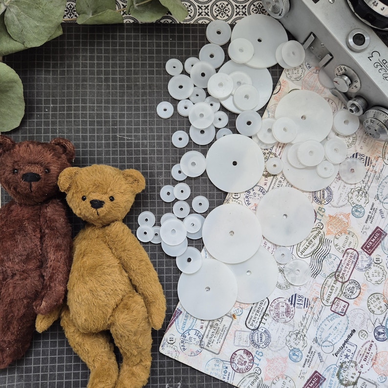 Op de afbeelding: Twee handgemaakte teddyberen, &eacute;&eacute;n bruin en &eacute;&eacute;n bruin, naast een verzameling witte plastic schijven met gaten. Een vintage camera en papier met patronen en stempels zijn ook zichtbaar.