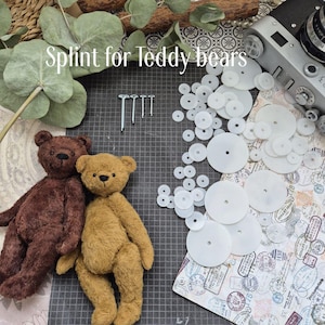 May include: Two teddy bears, one brown and one tan, sit beside a collection of white splints and fasteners. The text "Splint for teddy bears" is written across the top of the image. A vintage camera and greenery are also visible.