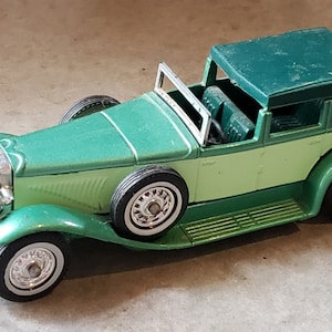 May include: A vintage-style toy car in shades of green, with a convertible top. The car features white-walled tires and chrome detailing on the front grill and headlights. The car is approximately 8 inches long.
