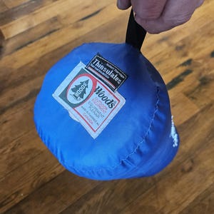 Puede incluir: Una bolsa azul de equipo para exteriores Woods con un asa negra. La bolsa tiene una etiqueta Thinsulate y una etiqueta Woods con texto en inglés y francés. Hecho en Canadá.