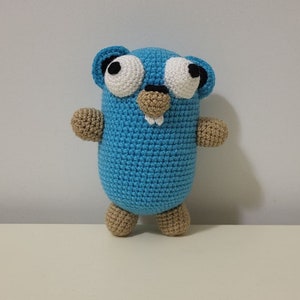 Può includere: Un giocattolo all'uncinetto blu fatto a mano a forma di personaggio dei cartoni animati con grandi occhi bianchi, piccole orecchie e muso e arti color beige. Il giocattolo ha un corpo arrotondato ed è in piedi.