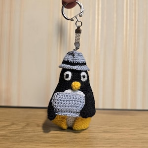 May include: A crocheted penguin keychain featuring a black body, a light gray belly, and yellow feet. The penguin wears a gray fedora-style hat and has a small yellow beak with white and black eyes. It is attached to a silver metal keyring.