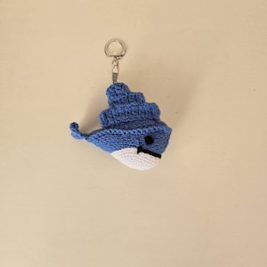 Puede incluir: Un llavero de ballena de ganchillo azul y blanco hecho a mano. La ballena tiene una cara sonriente con un ojo y una boca negros. El llavero está unido a un anillo de metal plateado.
