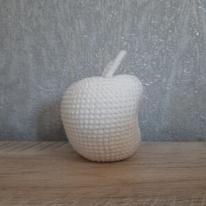 Op de afbeelding: Een witte gehaakte appel met een steel. De appel ligt op een houten oppervlak.