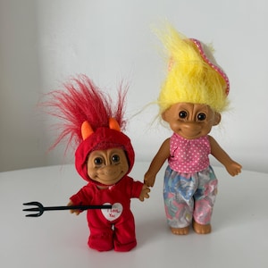 May include: Two troll dolls standing side by side. The left doll has red hair, orange horns, and a red outfit with a white heart featuring the text I Love You, holding a black plastic pitchfork. The right doll has yellow hair and wears a pink polka-dot top.