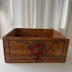Peut inclure: Une caisse en bois rectangulaire avec une finition marron naturel. Le panneau avant présente un texte noir indiquant 60 LBS. NET WGT. et CHURCH & DWIGHT CO. INC. NEW YORK, N.Y. Un logo circulaire affiche ARM & HAMMER SODA avec une icône de bras.