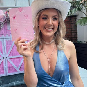 May include: A woman wearing a white cowboy hat and a light blue sparkly dress holds a pink card with a list of cocktails. The card has the text "Let's go girls" at the top.