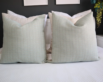 May include: Two square throw pillows featuring a light sage green fabric with a subtle, repeating vertical wavy line pattern. Each pillow is positioned in front of a plain, solid off-white pillow, creating a layered look on a white bedspread.
