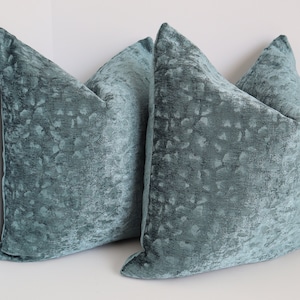 May include: Two teal blue velvet throw pillows with a subtle textured pattern. The pillows are square and have a simple design.