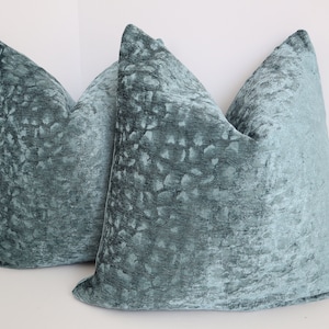 May include: Two teal blue velvet throw pillows with a subtle textured pattern. The pillows are square and have a simple design.