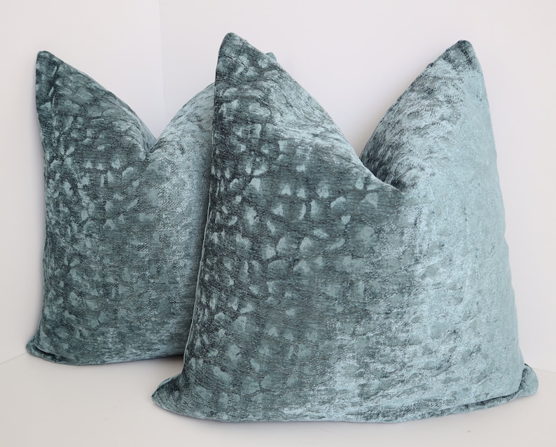 May include: Two teal blue velvet throw pillows with a subtle textured pattern. The pillows are square and have a simple design.
