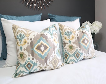 Puede incluir: Dos almohadas decorativas con un patrón geométrico en tonos marrones, azules y amarillos. Las almohadas están sobre una colcha blanca.