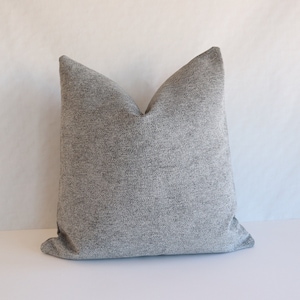 May include: A square, gray decorative pillow with a textured fabric. The pillow has a simple design and is likely used for home decor. The pillow is on a white surface.