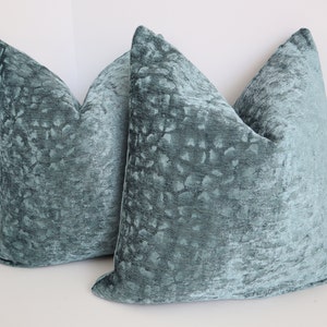 May include: Two teal blue velvet throw pillows with a subtle textured pattern. The pillows are square and have a simple design.