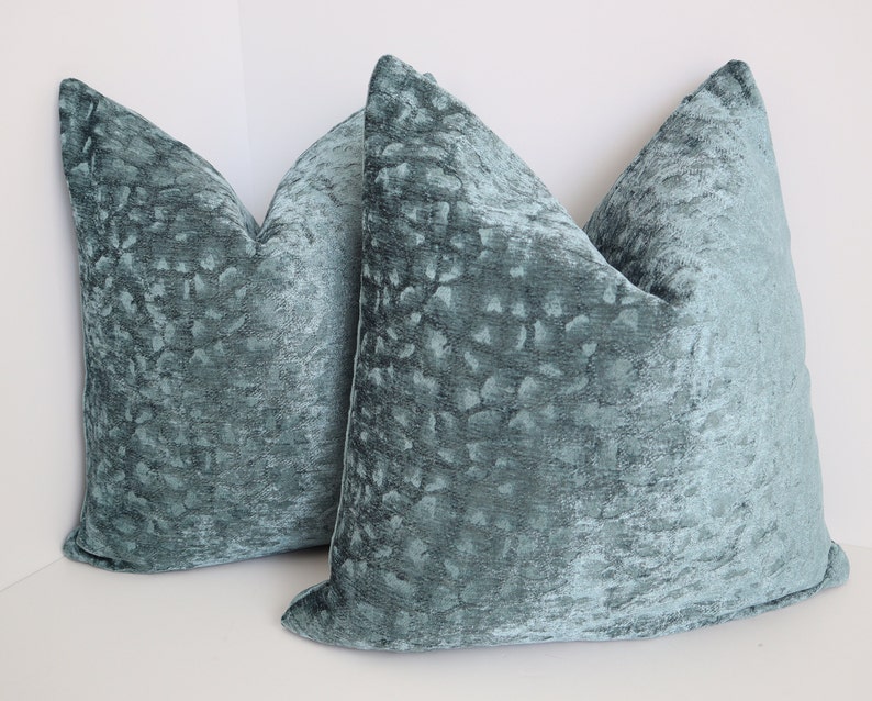 May include: Two teal blue velvet throw pillows with a subtle textured pattern. The pillows are square and have a simple design.