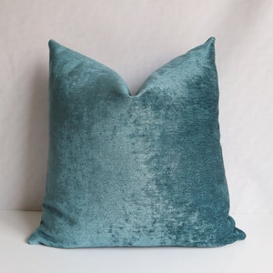 Puede incluir: Una almohada de terciopelo azul turquesa con una superficie suave y texturizada. La almohada es de forma cuadrada y tiene un dise&ntilde;o simple.