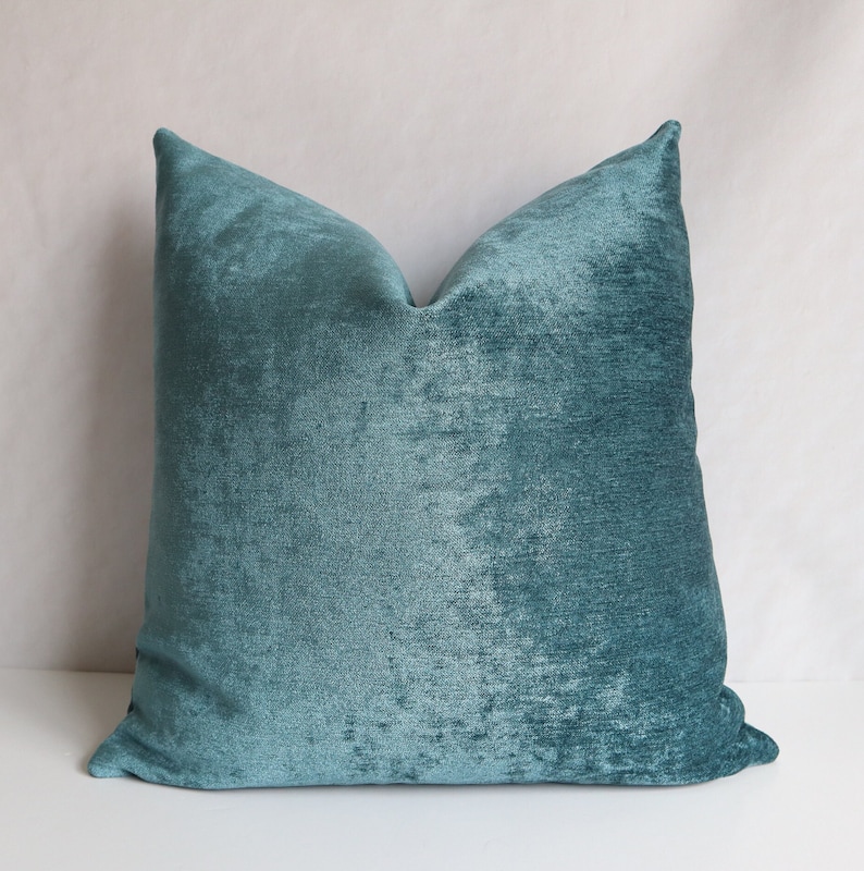 Puede incluir: Una almohada de terciopelo azul turquesa con una superficie suave y texturizada. La almohada es de forma cuadrada y tiene un dise&ntilde;o simple.