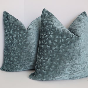 May include: Two teal blue velvet throw pillows with a textured, embossed pattern. The pillows are arranged in a corner, with one pillow partially overlapping the other.