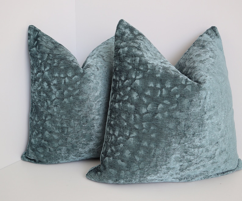 May include: Two teal blue velvet throw pillows with a textured, embossed pattern. The pillows are arranged in a corner, with one pillow partially overlapping the other.