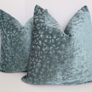 May include: Two teal blue velvet throw pillows with a subtle textured pattern. The pillows are square shaped and have a soft, plush feel.