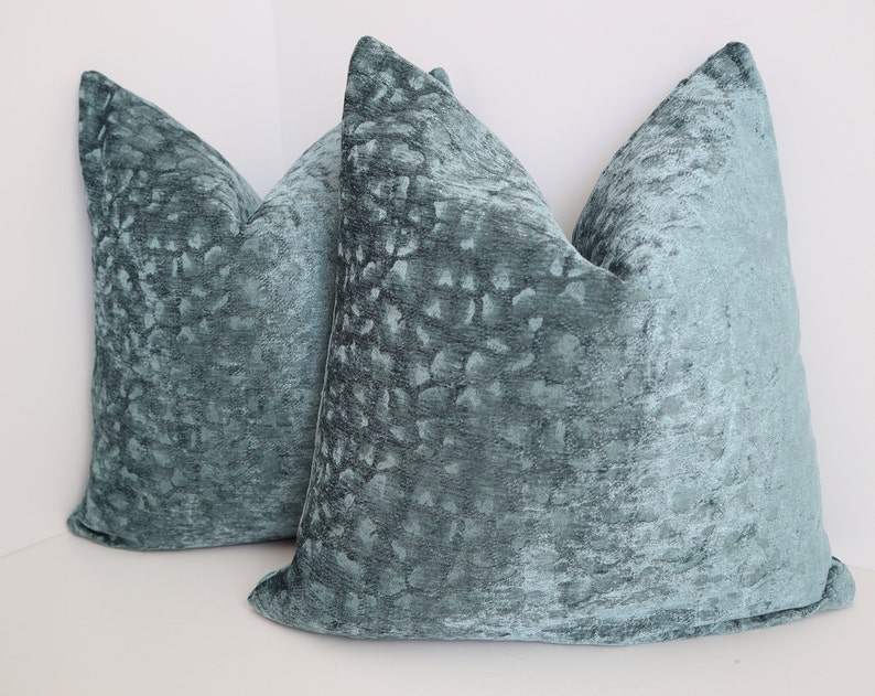 May include: Two teal blue velvet throw pillows with a subtle textured pattern. The pillows are square shaped and have a soft, plush feel.