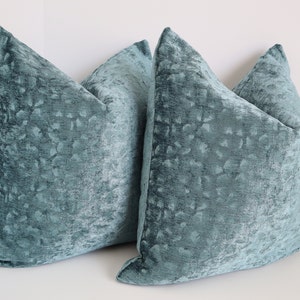 May include: Three teal blue velvet throw pillows with a subtle textured pattern. The pillows are arranged in a corner, with two pillows stacked on top of each other and the third pillow in front.