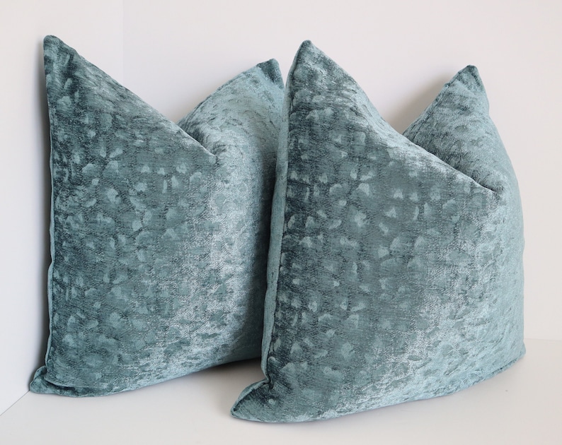 May include: Three teal blue velvet throw pillows with a subtle textured pattern. The pillows are arranged in a corner, with two pillows stacked on top of each other and the third pillow in front.