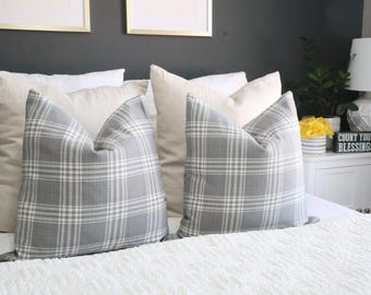 May include: Two gray and white plaid throw pillows rest on a white bed, with several solid-colored pillows behind them. The pillows are arranged on a bed with a white comforter. The room has a neutral color scheme.