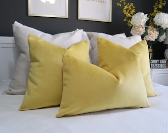 May include: A collection of three square and rectangular throw pillows featuring a smooth, solid yellow velvet fabric. These pillows are arranged on a white bedspread, layered in front of larger, neutral-toned beige and white square pillows.