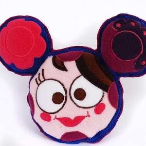 May include: A plush, cartoon-style pillow in the shape of a face with large, round eyes and a smiling mouth. The pillow features a light pink face with dark pink and brown accents, red lips, and blue trim. The ears are red and purple.