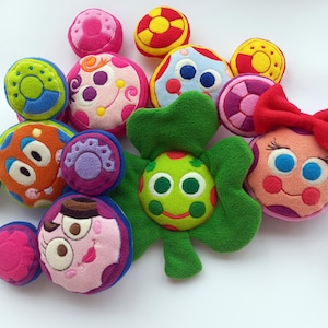 May include: A selection of brightly coloured plush toys, including balls and flower-like shapes. The toys feature various designs, including faces and circles, in shades of pink, green, orange, blue, and yellow. Some have embroidered details.