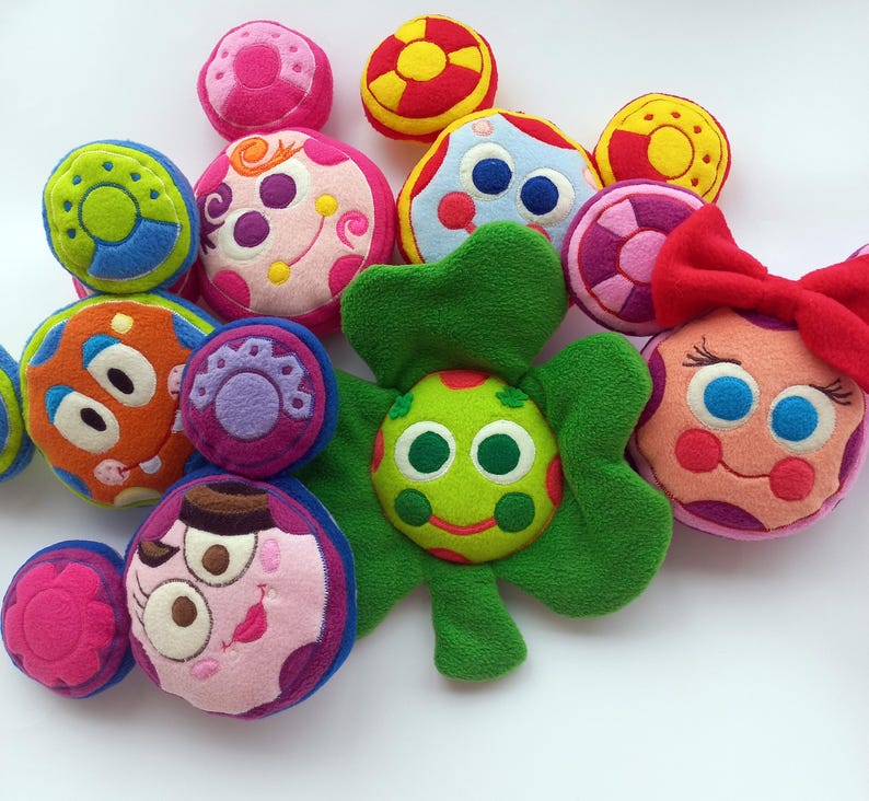 May include: A selection of brightly coloured plush toys, including balls and flower-like shapes. The toys feature various designs, including faces and circles, in shades of pink, green, orange, blue, and yellow. Some have embroidered details.