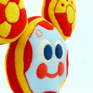 May include: A soft toy featuring a cartoon face and three circular elements. The face is light blue with large blue eyes, red cheeks, and a red smile. The circular shapes are red and yellow, with a striped design.
