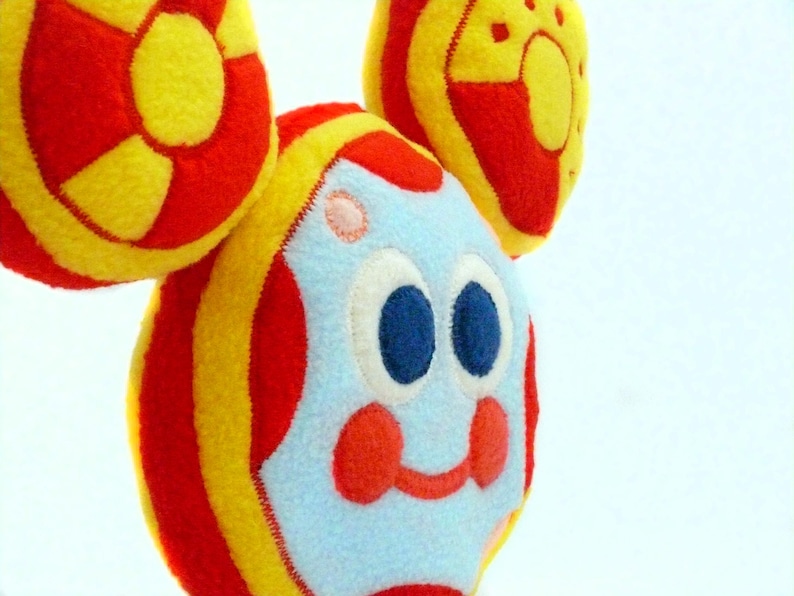 May include: A soft toy featuring a cartoon face and three circular elements. The face is light blue with large blue eyes, red cheeks, and a red smile. The circular shapes are red and yellow, with a striped design.