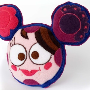 May include: A plush, cartoon-style pillow in the shape of a face with large ears. The pillow features a pink and blue color scheme with brown hair, large eyes, and a smiling mouth. The ears have a flower-like design.