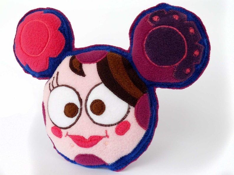 May include: A plush, cartoon-style pillow in the shape of a face with large ears. The pillow features a pink and blue color scheme with brown hair, large eyes, and a smiling mouth. The ears have a flower-like design.
