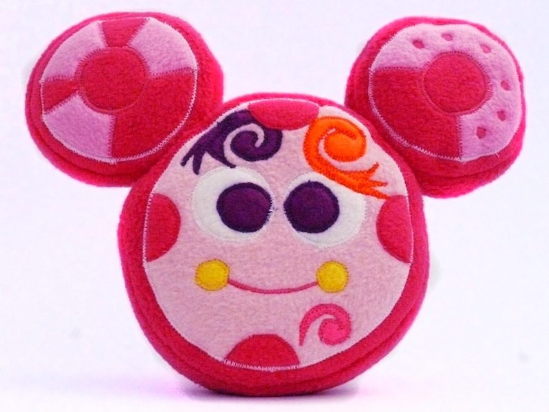 May include: A soft toy resembling a cartoon character's face, predominantly pink and light pink. It has large purple eyes, yellow cheeks, and orange and purple decorative details. The circular ears echo the face's design.