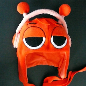May include: Orange fleece hat with large, sleepy eyes, a white headband, and two orange ball-shaped antennae. The hat has a face design with black and white details, and ear flaps with long ties.